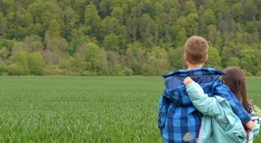 Two children looking out over a green field with a forest in the background.