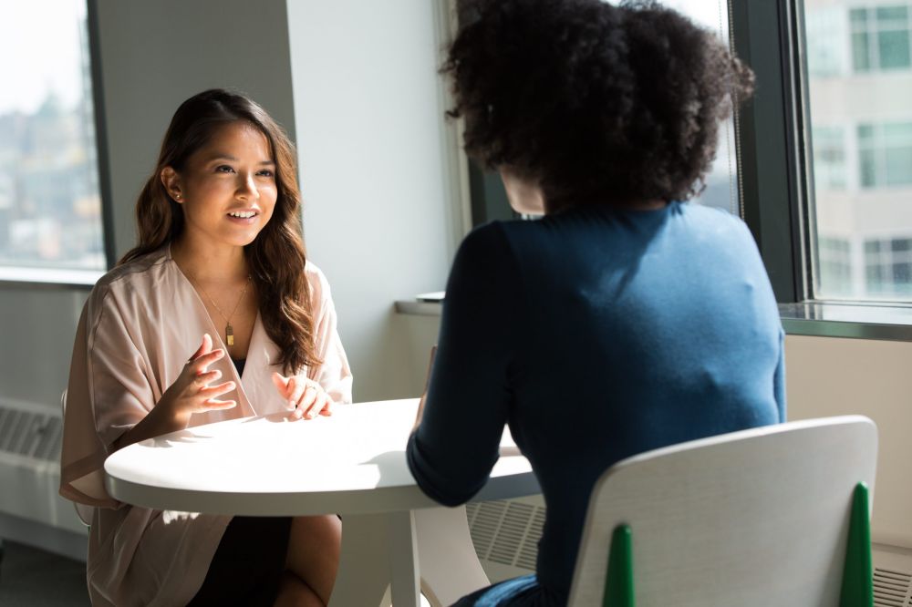 Two individuals in a discussion at a round table, indoors with city view outside