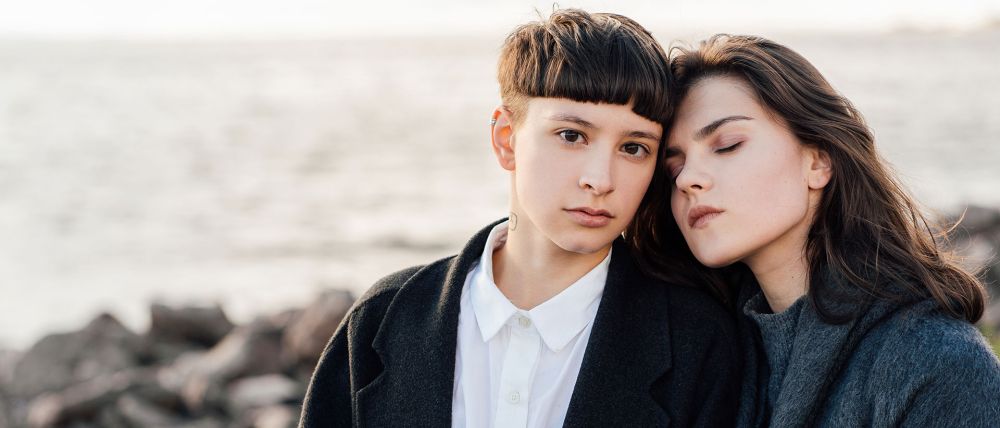 Two women resting heads together by a rocky shoreline, displaying a close and affectionate bond.