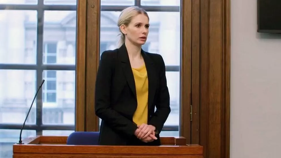 Person in a black blazer and yellow top standing at a wooden podium in a room with windows.