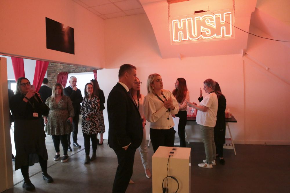 People standing inside a room with a HUSH sign on the wall.