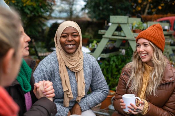 Three people sitting outdoors, one holding a white mug and two with clasped hands.