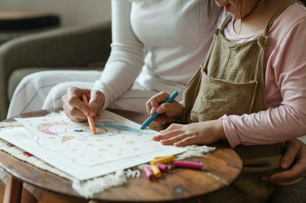 A child and an adult color a unicorn drawing with crayons at a wooden table. The child wears a light pink shirt and tan overalls, while the adult wears white clothes.