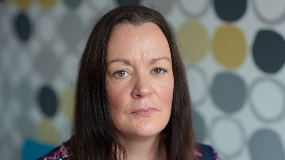Close-up of Kate Wallace with long brown hair, neutral expression, and a patterned background.